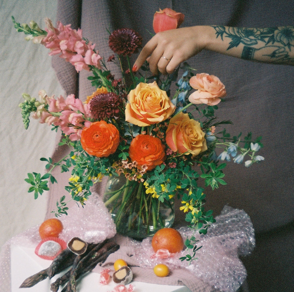 a tattooed hand adjusting a vibrant floral bouquet on a citrus-themed thread created by Bagel's Florals in NM