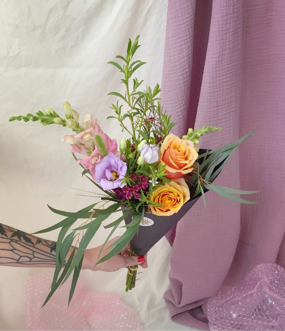 white tattooed hand holding a bouquet of orange, purple, and pink flowers and greenery